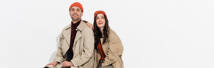 panoramic shot of stylish woman in trench coat looking up near trendy man in beanie hat on white