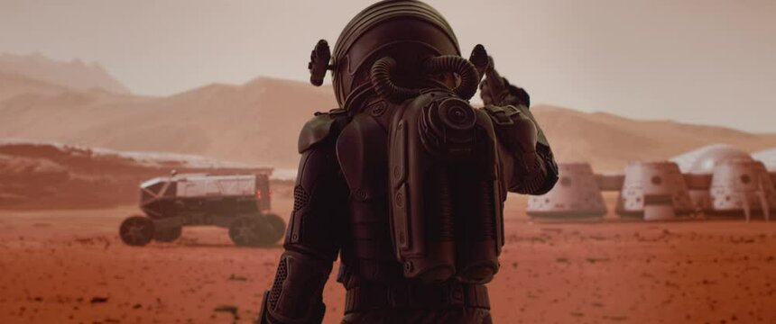 HANDHELD TRACKING back view of astronaut wearing space suit walking on a surface of a red planet. Martian base and rover in the background. Mars colonization concept