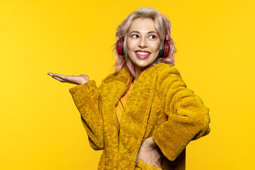 Funny blonde girl in headphones and a colored fur coat on a yellow background holds an outstretched...