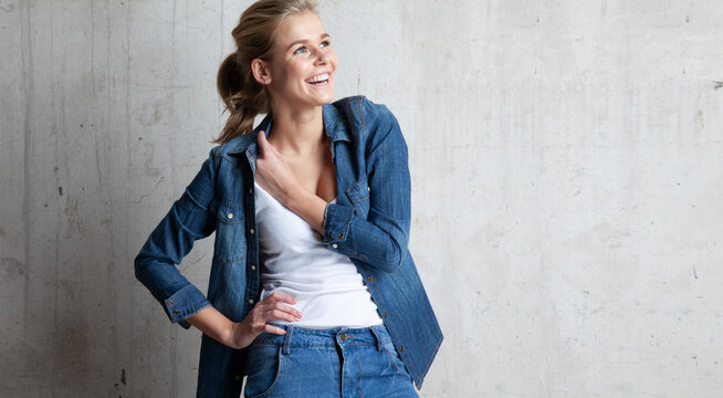 Portrait of perfect model posing in studio in casual jeans jacket and white t-shirt.Attractive happy girl with sincere smile. Concrete wall on background. Copy space in left side
