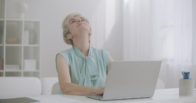 retiree woman with osteochondrosis is stretching hands and neck during work with laptop, sitting at table and typing on keyboard