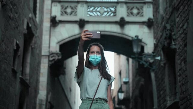 Beautiful long-haired brunette in protective mask makes selfie under the arch. Young attractive woman taking pictures in a beautiful city. COVID-19 precaution. Stay safe. Tourist with mobile phone