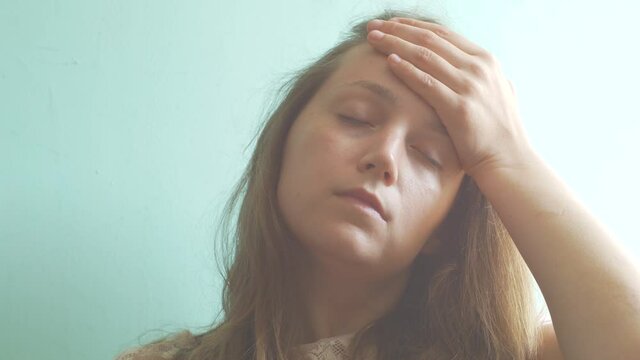 Stressed brown hair woman with headache on pastel mint background. Tired young lady without make-up feel pain, massaging temples. Hard to wake up, morning concept. PMS premenstrual syndrome problems.