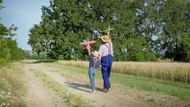 parenting, elderly happy grandfather together with his beloved grandson in straw hats have fun by launching toy planes walking along path backdrop of green trees outside city