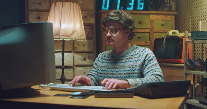 Caucasian male nerd in glasses with mustache sitting at desk in retro room and working on computer. Man programist typing on keyboard while studying. Vintage style of 90's. Hacker from 80's.