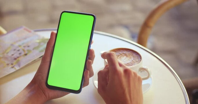 Crop view of female person holding smartphone and scrolling on green screen while sitting at table with coffee and map on it. Concept of mockup and chroma key.