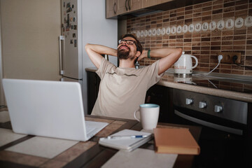 Smiling hipster freelancer holding hands behind head sitting at office desk behind laptop.