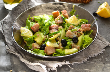 Salad with lettuce, fish, blueberries, lemon and avocado. Healthy lunch or brunch. Dark background. Linen gray napkin. Salad in a black glass plate. Backlight. Fish bowl with avocado.