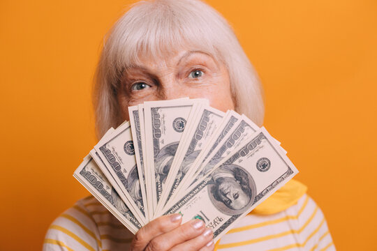 image of cheerful elderly woman with gray eyes and white short hair wearing yellow cravat, striped t-shirt and holding a fan of dollars. Woman isolated over orange background. Looking at camera.
