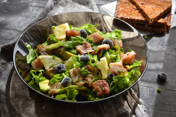 Salad with lettuce, fish, blueberries, lemon and avocado. Healthy lunch or brunch. Dark background. Linen gray napkin. Salad in a black glass plate. Backlight. Fish bowl with avocado.