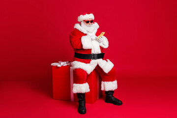 Full length photo of retired old man white beard hold telephone sit large box what chatting elf friend wear x-mas santa costume sunglass headwear leather boot isolated red color background