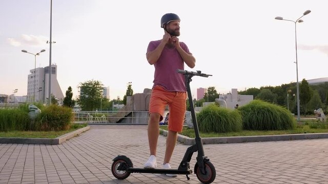 A young modern bearded man in a protective helmet rides an electric scooter in the Park in the summer.