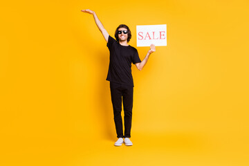 Full legth photo of young man stand hold poster sale raise hand empty space wear black t-shirt pants white sneakers sunglass isolated yellow color background