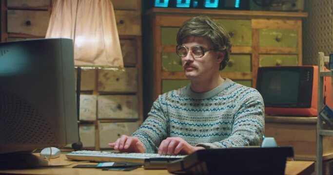 Portrait of Caucasian male nerd in glasses with mustache sitting at desk in retro room, smiling and working on computer. Funny man programist looking at camera and typing on keyboard. Vintage style.