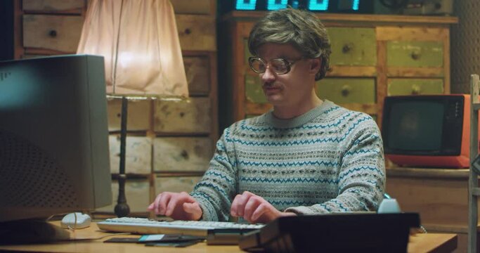 Portrait of Caucasian male nerd in glasses with mustache sitting at desk in retro room, smiling and working on computer. Man programist typing on keyboard and giving thumb up to camera. Vintage style.