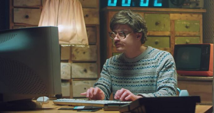 Portrait of Caucasian male goofy nerd in glasses with mustache sitting at desk in retro room and working on computer. Funny man programist looking at camera and typing on keyboard. Vintage style.