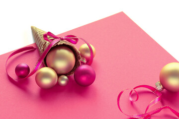 Golden waffle ice cream cone with Xmas gold balls, stars and ribbons on pink paper. Text Merry Christmas. Xmas decor arrangement isolated on white.