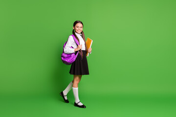 Photo portrait of schoolgirl going forward holding books in hand wearing violet backpack on shoulder isolated on vivid green colored background with copyspace - Powered by Adobe