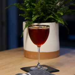 Elysian Cocktail in Nick & Nora gold rimmed glass on wood table with plant in background with a deep red color