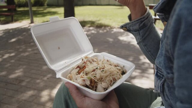 Woman Eating Rice Noodles With Vegetables In A Styrofoam Lunch Box Using Chopsticks While Sitting In The Empty Park - close up