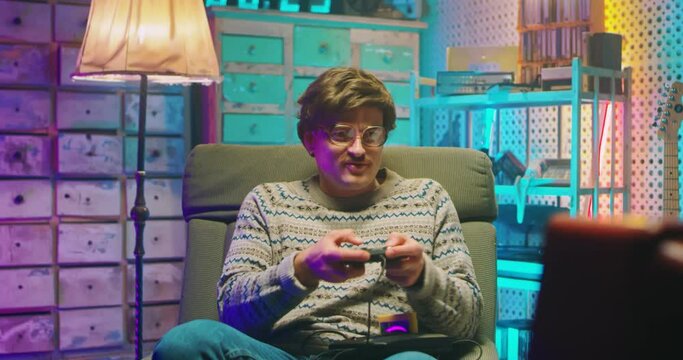 Caucasian man nerd in glasses and with mustache sitting in front of vintage TV monitor and playing videogame with joystick. Male silly goofy loosing in game. Retro style of 80's. Gamer loser from 90's