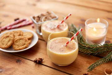 christmas and seasonal drinks concept - glasses of eggnog with oatmeal cookies, candy canes, sugar, fir tree branches and candle burning on wooden background