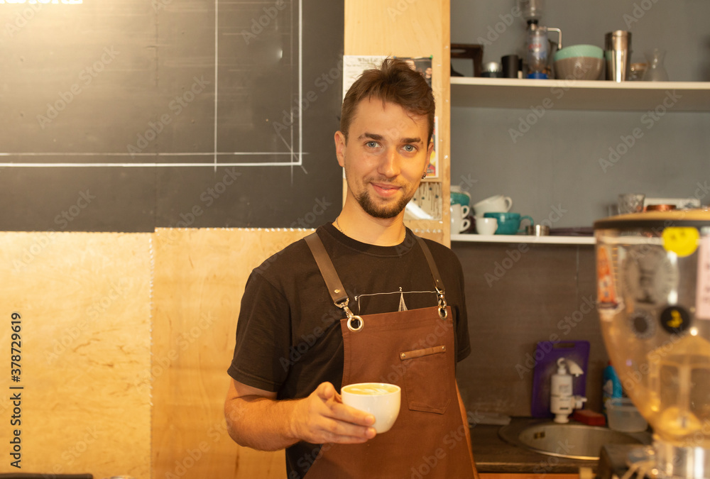 Poster handsome barista in apron holding a cup of coffee and smiling while working at the bar counter in ca