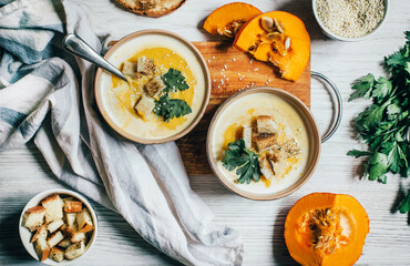 two plates of pumpkin soup next to greens pumpkin and bread croutons on a white wooden table top view close up
