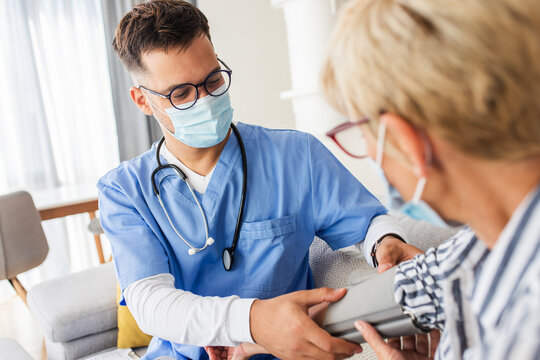 Male Nurse Measures Blood Pressure To Senior Woman With Mask While Being In A Home Visit.