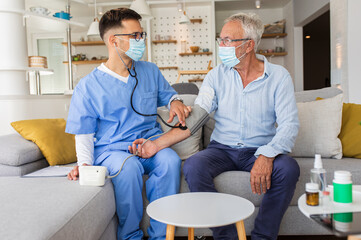 Fototapeta premium Male nurse measures blood pressure to senior man with mask while being in a home visit. 