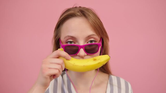 Woman wearing pink sunglasses makes smiling banana-shaped lips isolated on pink background