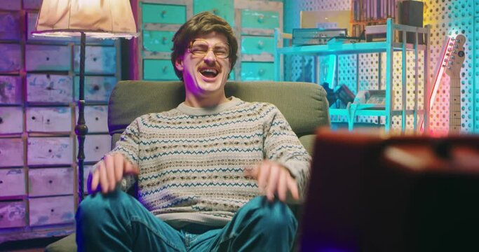 Caucasian funny young man nerd in glasses and with mustache sitting in retro style home and watching TV. Vintage television of 80's. Male laughing at comedy movie, film or program. Goofy guy of 90's.