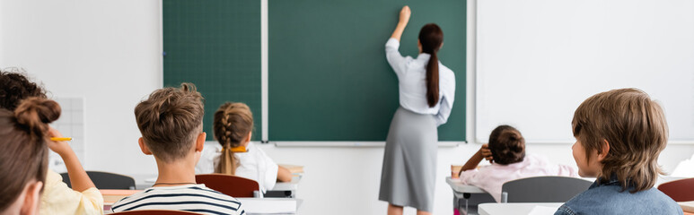 back view of teacher writing on chalkboard near multiethnic pupils during lesson, horizontal image - obrazy, fototapety, plakaty