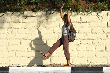 Young woman tourist with backpack behind her back walks cheerfully along curb stone by the road, waving her hands up on summer sunny day