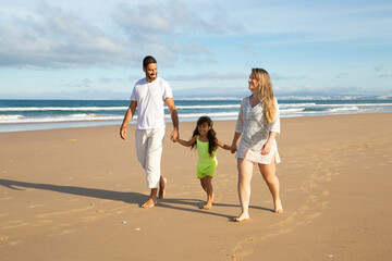Cheerful parents and kid in summer clothes walking on golden sand on beach, mom and dad holding girls hands. Full length. Family outdoor activities concept