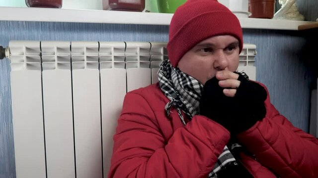 Male freeze at home and sits near house pipe heating system. White metal heating iron household radiator inside cold room near wall. Budget in crisis, feels cold temperature. Warming hands in gloves