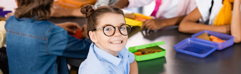 selective focus of excited schoolgirl in eyeglasses looking at camera near classmates and lunch...