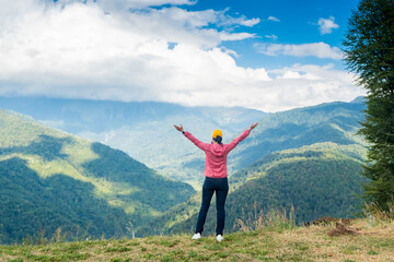 Happy young woman stands on a cliff with raised arms and looks at the mountains in front