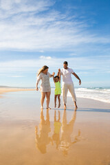 Happy family couple and little girl enjoying walking and activities on beach, kid holding parents hands, jumping and hanging. Front view. Family outdoor activities concept