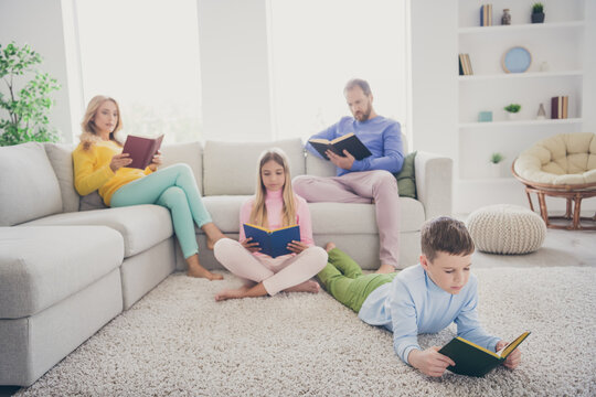 Full body photo of focused mom dad sit couch two small kids boy girl lying floor read book in house indoors