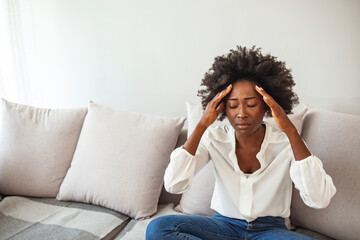 Shot of a young woman suffering from a headache. Young woman sitting on a couch, holding her head, having a strong headache. Sad girl suffering from headache. 