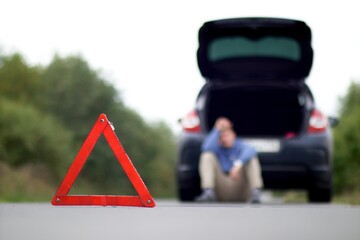 Accident on the road. Emergency warning triangle and a blurred man sitting near damaged car with open trunk