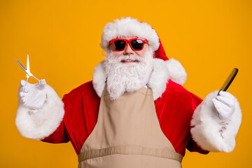 Close-up portrait of his he nice attractive cheerful cheery grey-haired Santa wearing apron holding in hands coiffure tools equipment isolated bright vivid shine vibrant yellow color background