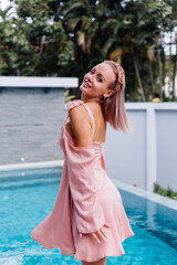 Happy caucasian calm stylish cute woman in pink summer dress and elegant brown headband standing on teracce at villa enjoying beautiful day alone.  