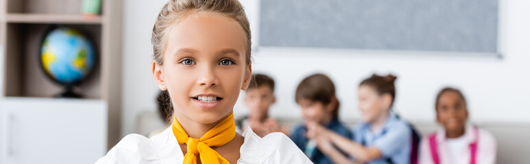 Panoramic crop of schoolgirl looking at camera with miltiethnic classmates at background