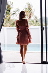 Happy caucasian calm stylish cute woman in pink summer dress and elegant brown headband standing on teracce at villa enjoying beautiful day alone.  