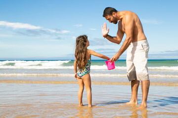 Handsome dad and little daughter picking shells with bucket on beach together, giving high five. Bright blue sea with waves and white foam in background. Family summer holidays concept