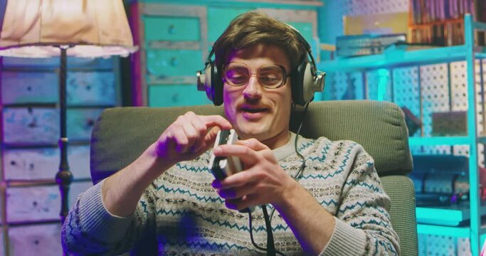 Young Caucasian man nerd in glasses, headphones and with mustache sitting in armchair in retro interior room and listening to music on vintage player. Male goofy imitating guitar play.