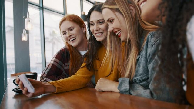 Female friends looking at the phone and smiling at the restaurant. Group of multi-ethnic women looking at pictures on smartphone and laughing.
