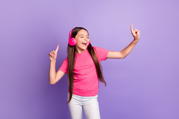 Fototapeta premium Photo portrait of singing smiling little girl listening to music with pink earphones singing pointing up with fingers on both hands isolated on purple color background